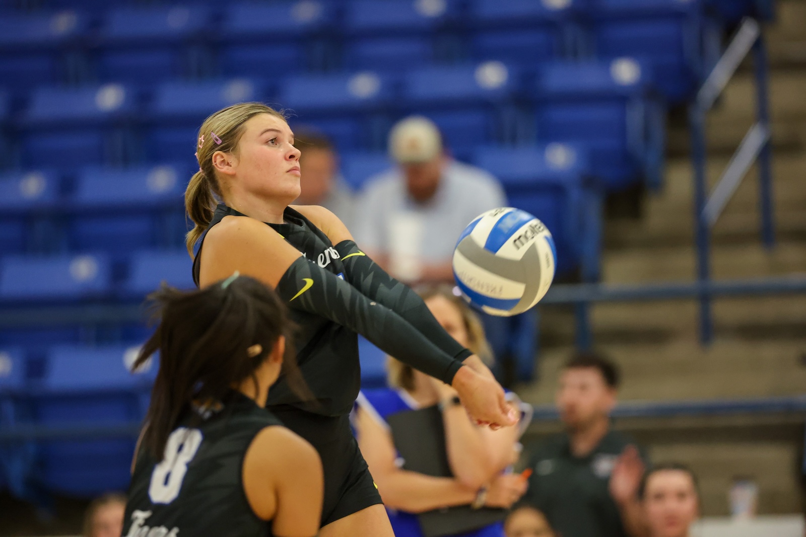 Seabolt Chase-12802 | Press Pass Sports Female volleyball player in black jersey stretches to pass ball during a indoors match, blue bleachers in the background.