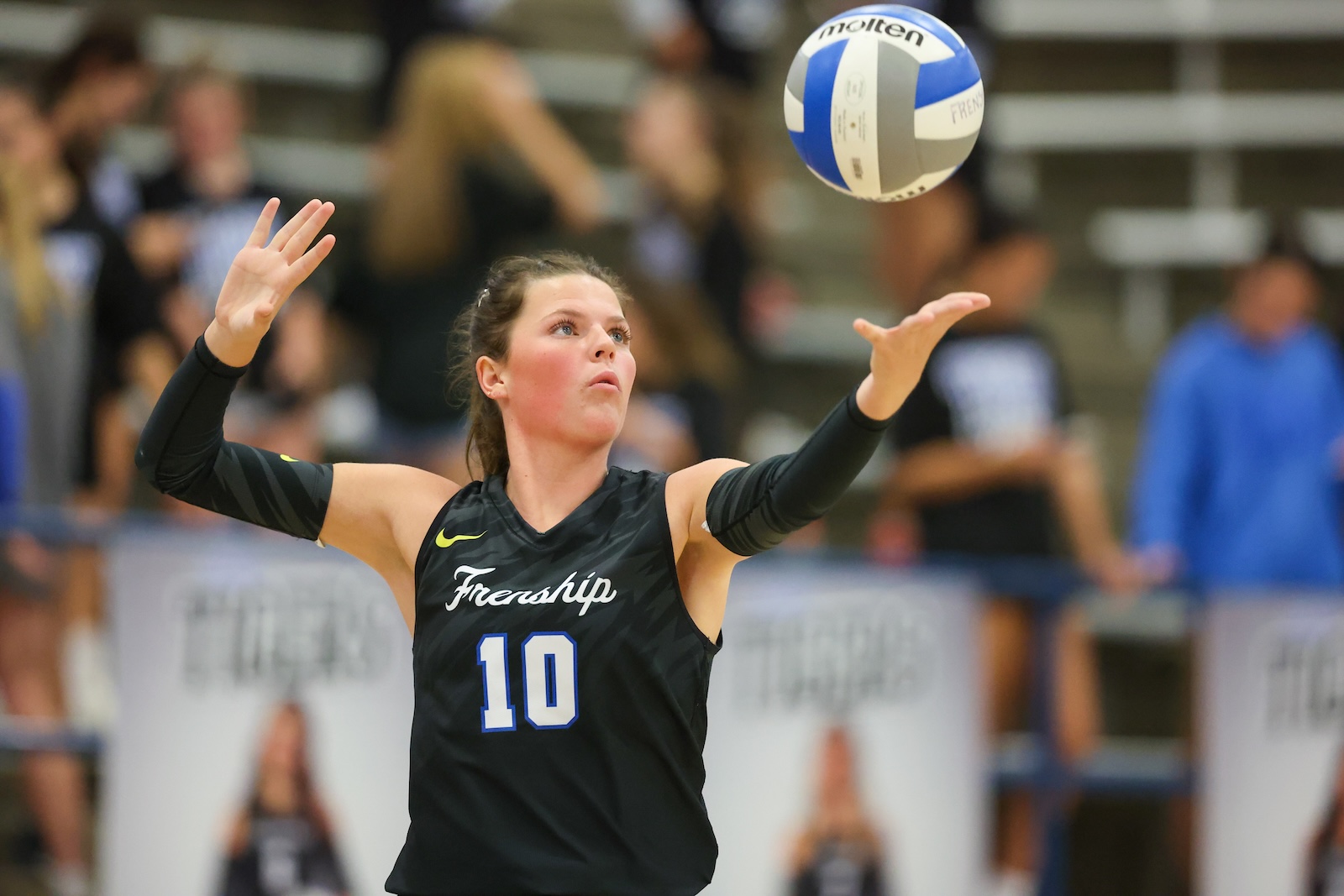 Seabolt Chase-12824 | Press Pass Sports Female volleyball player in a black jersey with number 10, arms raised to set a blue-and-white ball in a crowded gym.