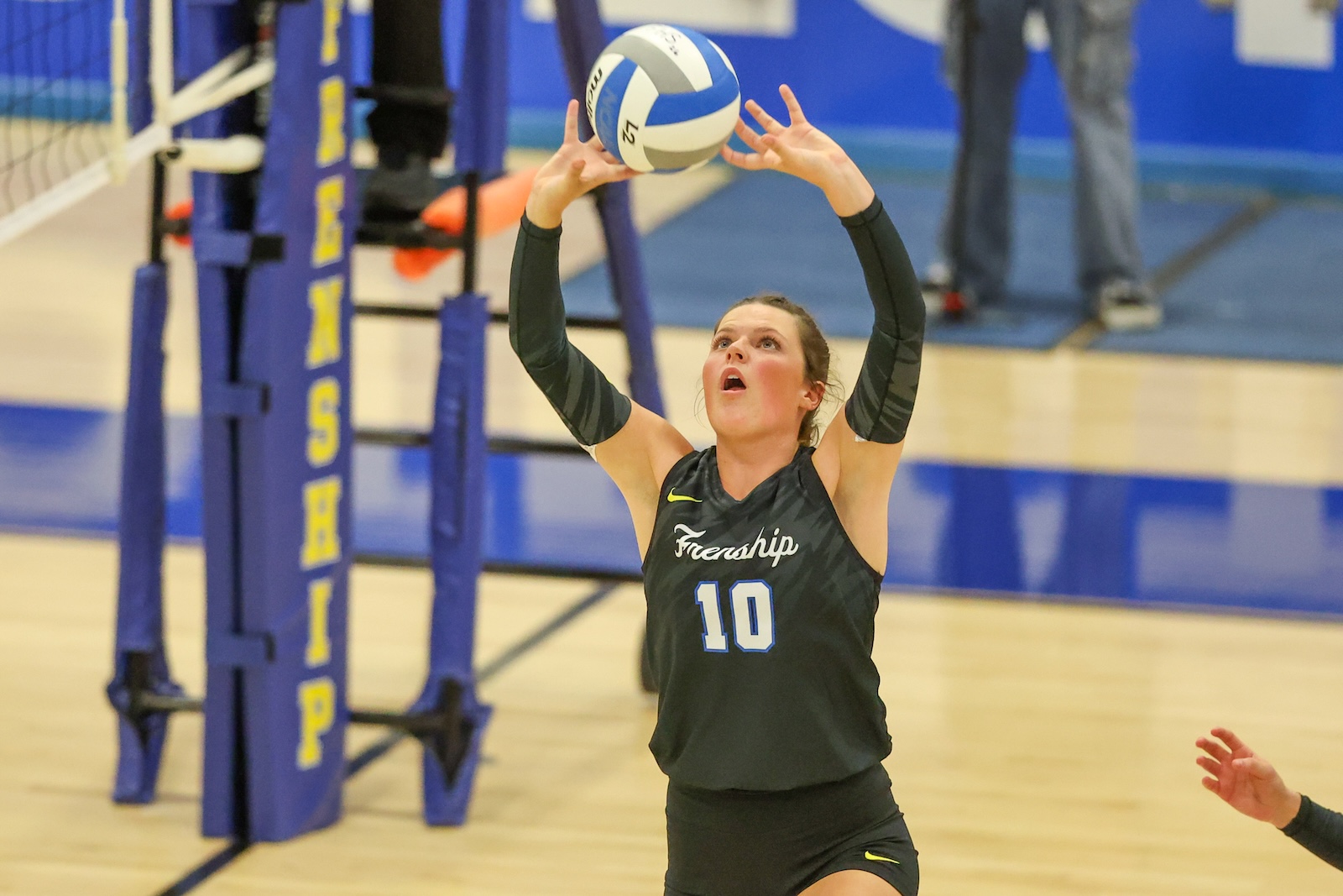 Seabolt Chase-12888 | Press Pass Sports Female volleyball player in a dark uniform (number 10) has arms raised to set a white-and-blue ball during a match on an indoor court in blue-and-gold surroundings.