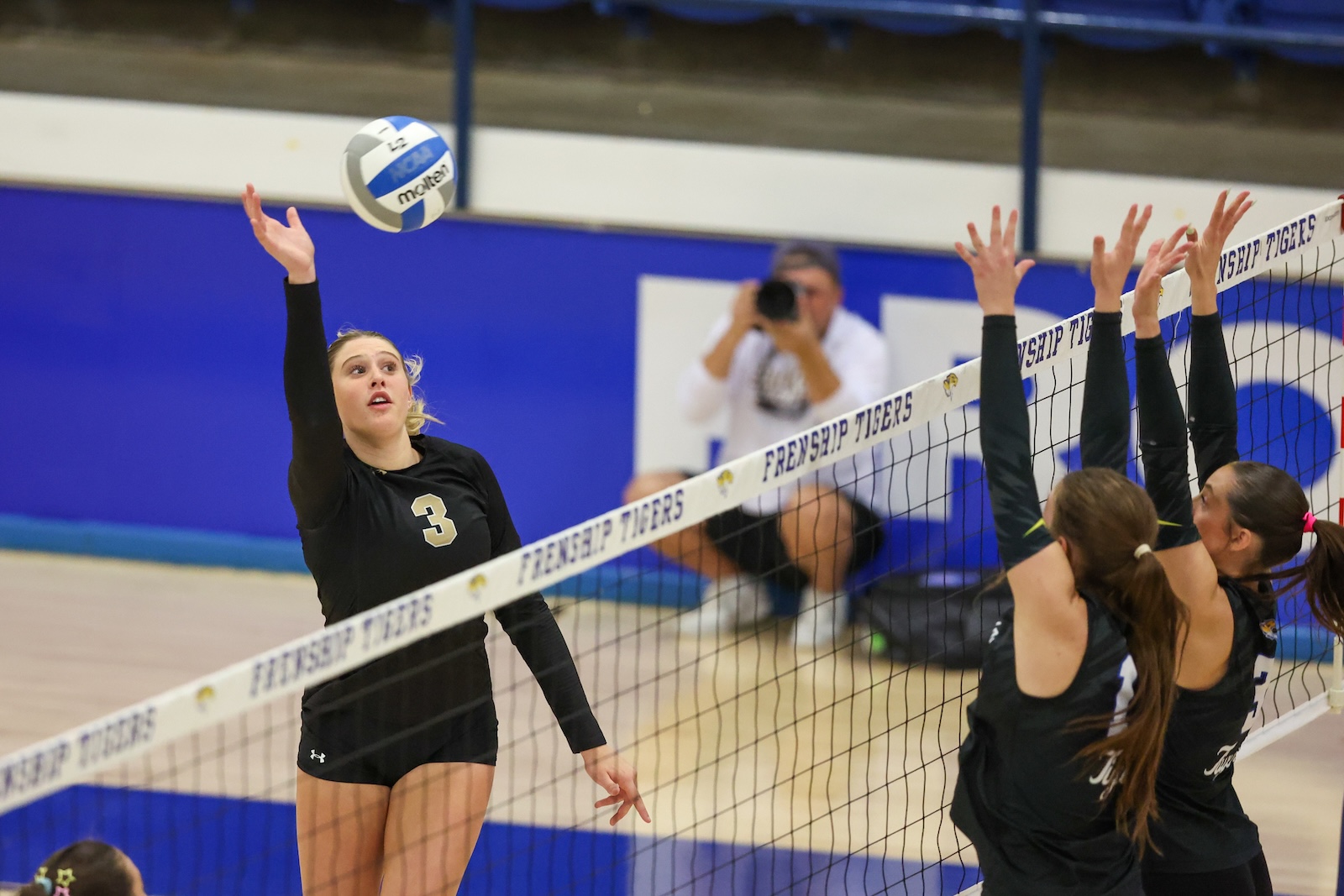 Seabolt Chase-12916 | Press Pass Sports Player in black with jersey number 3 spikes the ball over the net while teammates attempt a block.