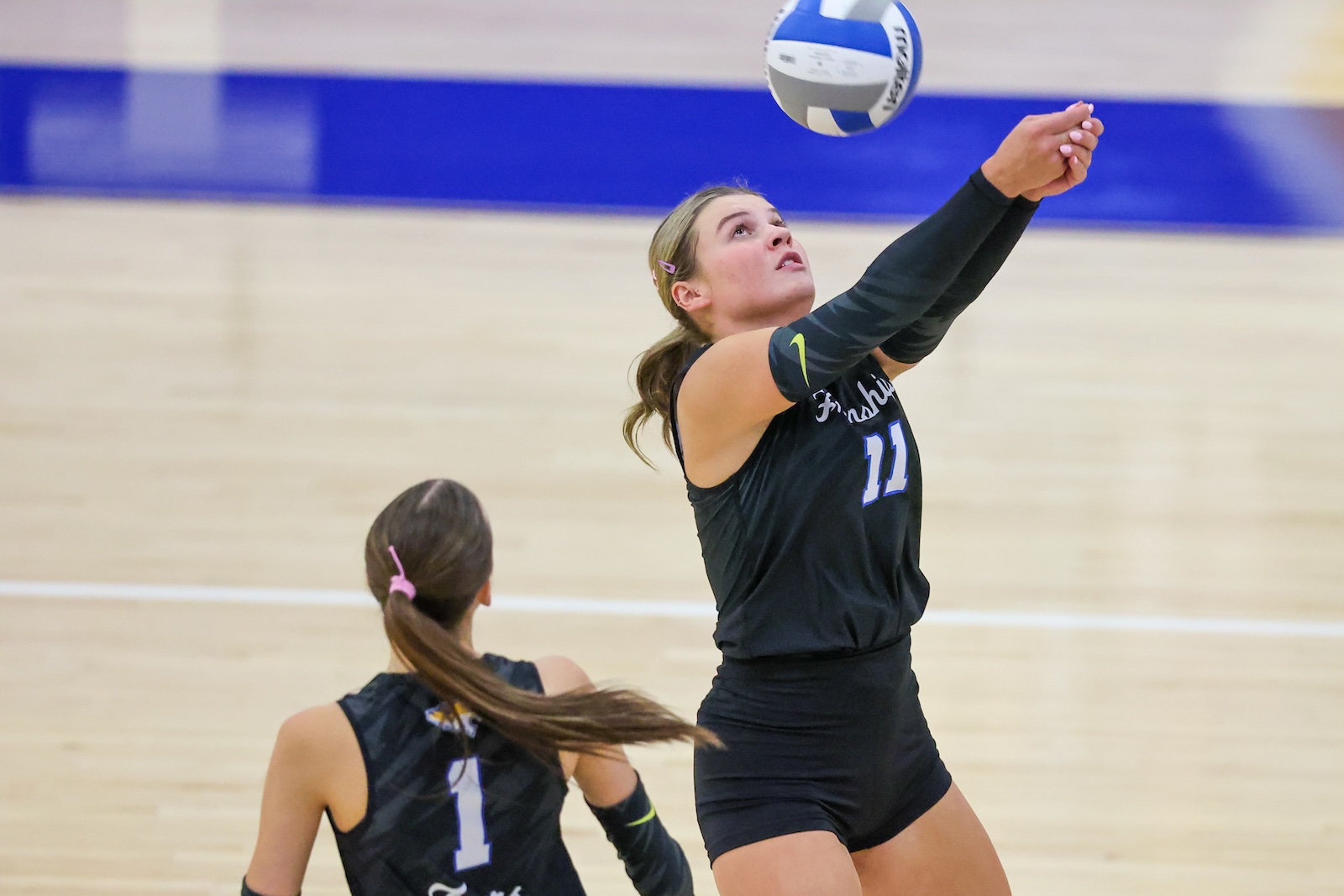 Seabolt Chase-12920 | Press Pass Sports Female volleyball player in black jersey #11 reaches up to hit a ball during a rally, another teammate in foreground watching inside a gym.