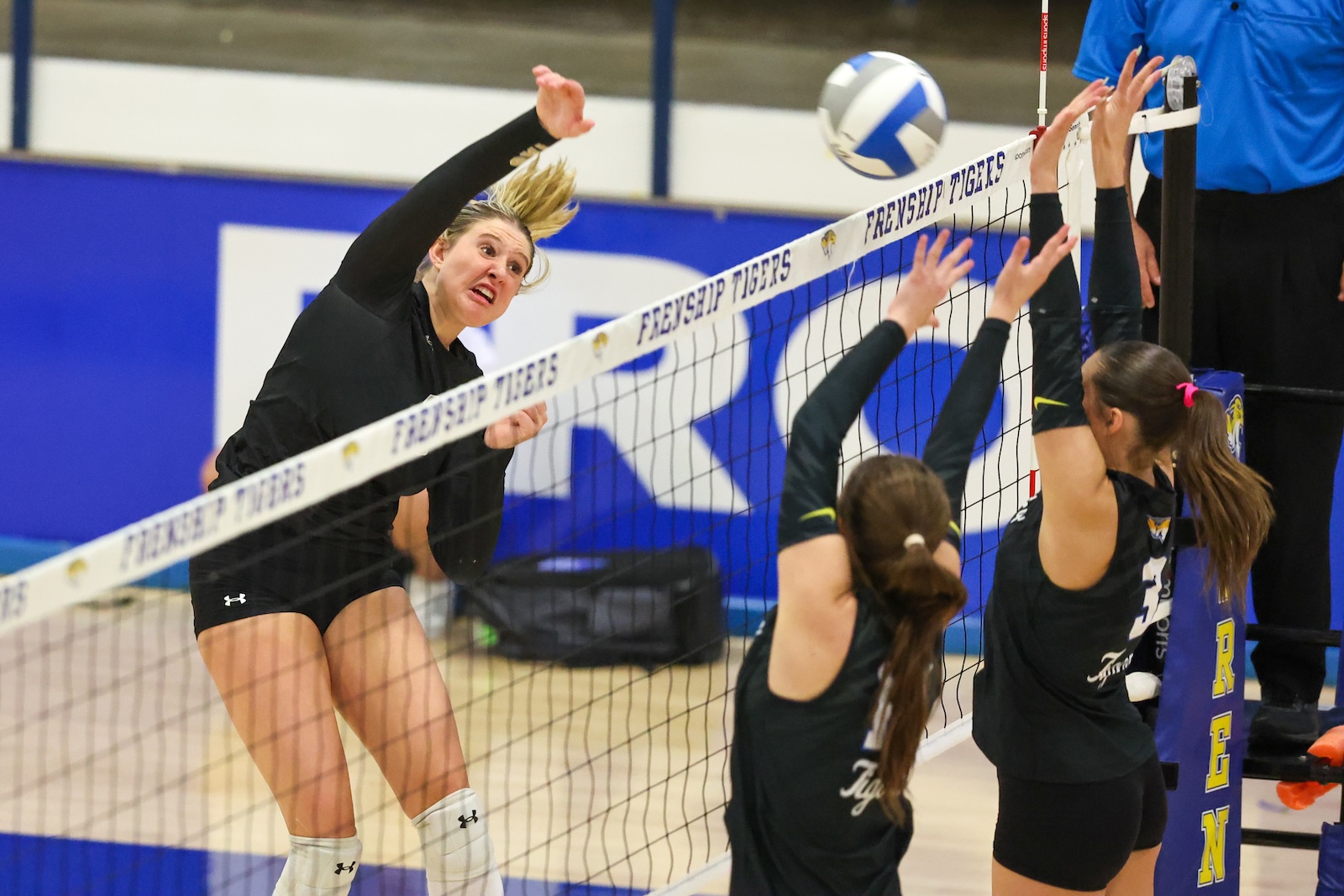 Seabolt Chase-12937 | Press Pass Sports Female volleyball player leaps to spike the ball over the net as two defenders jump to block.