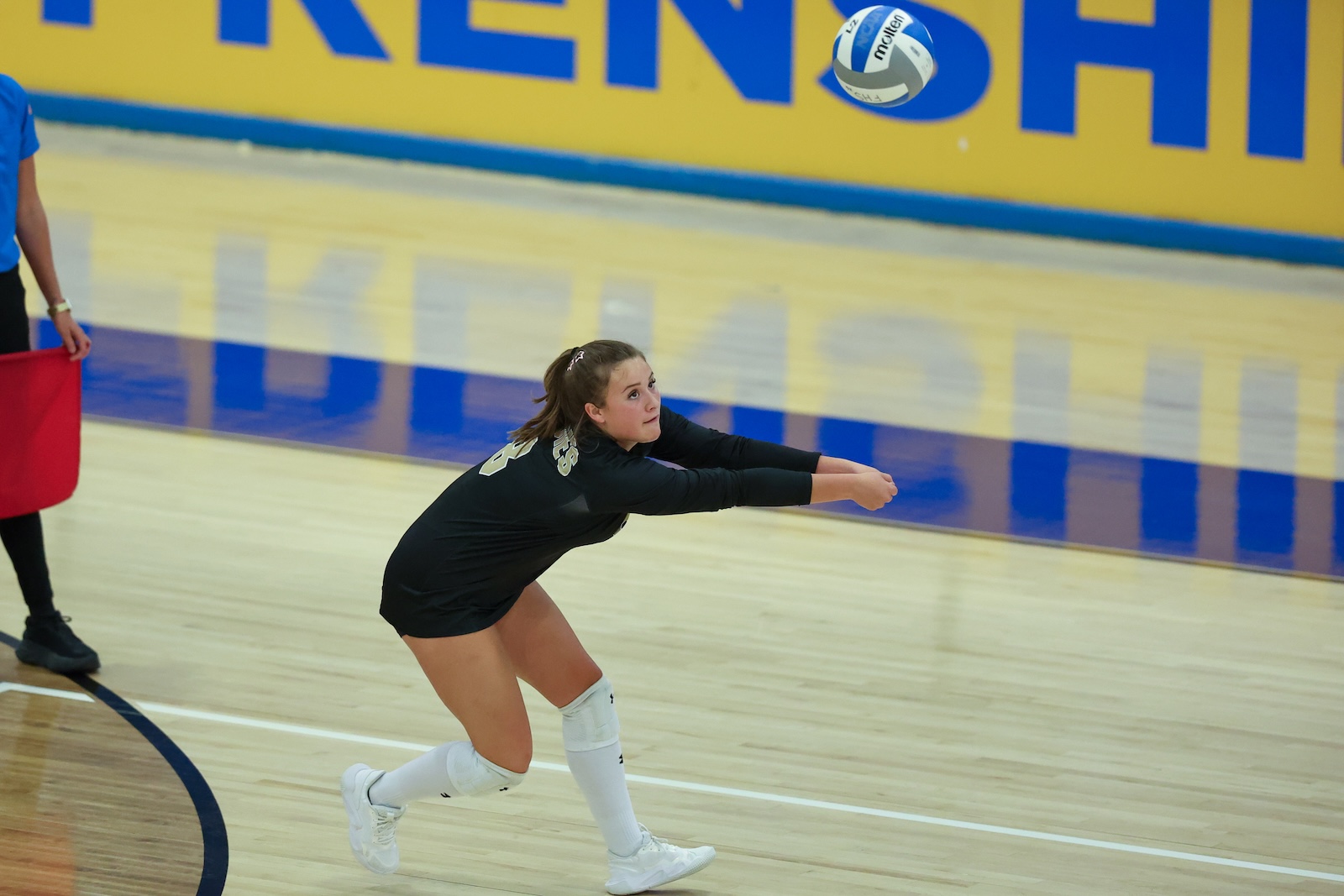 Seabolt Chase-13016 | Press Pass Sports Female volleyball player in black dives to save a ball on an indoor court; yellow wall background and blue lines visible.