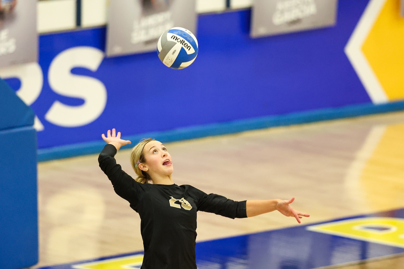 Seabolt Chase-13052 | Press Pass Sports Young female volleyball player in a black shirt reaches up toward a ball in midair inside a crowded gymnasium, ready to hit.