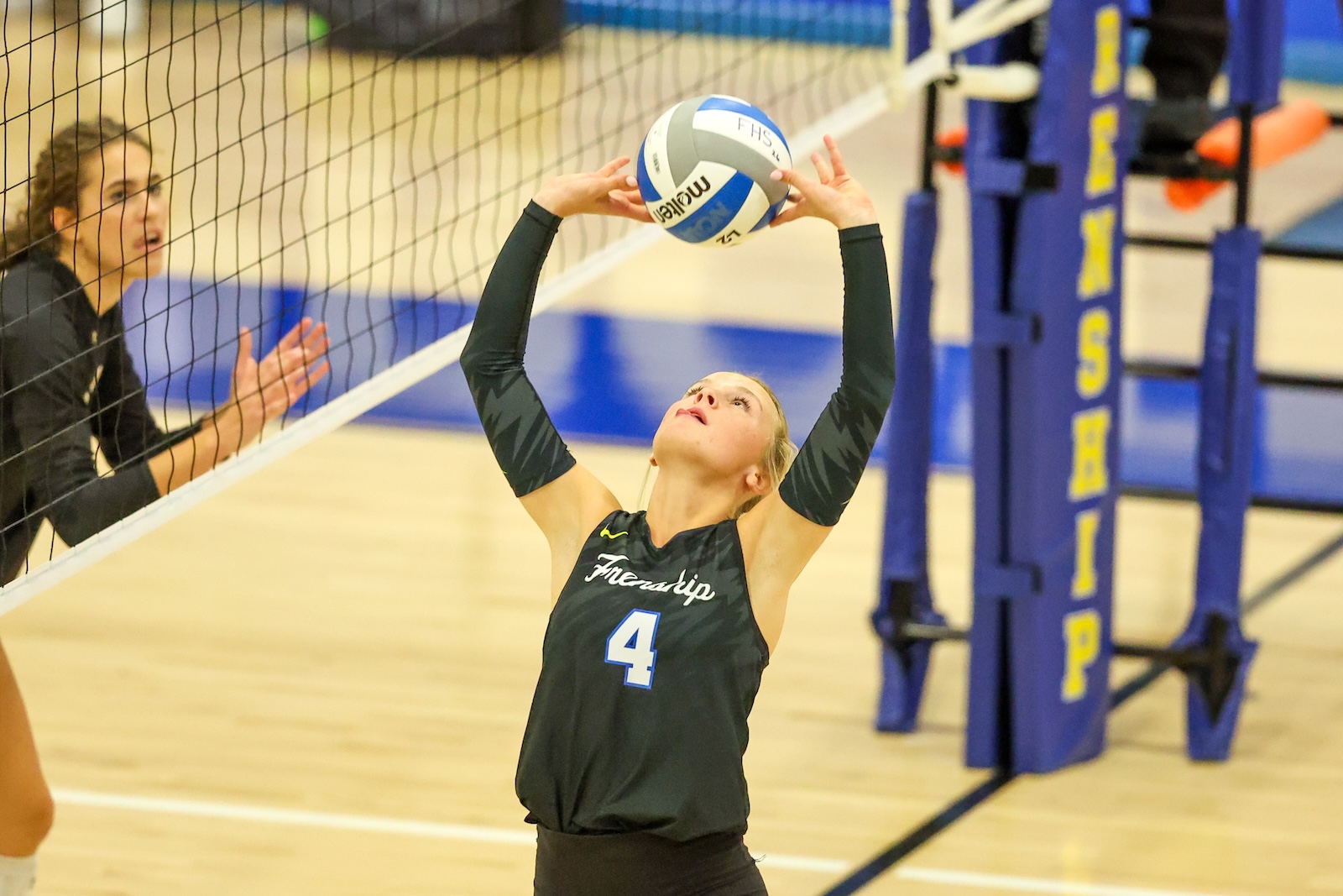 Seabolt Chase-13074 | Press Pass Sports Female volleyball player with jersey number 4 raises arms to set the ball at the net during a match, teammate clapping on the left.