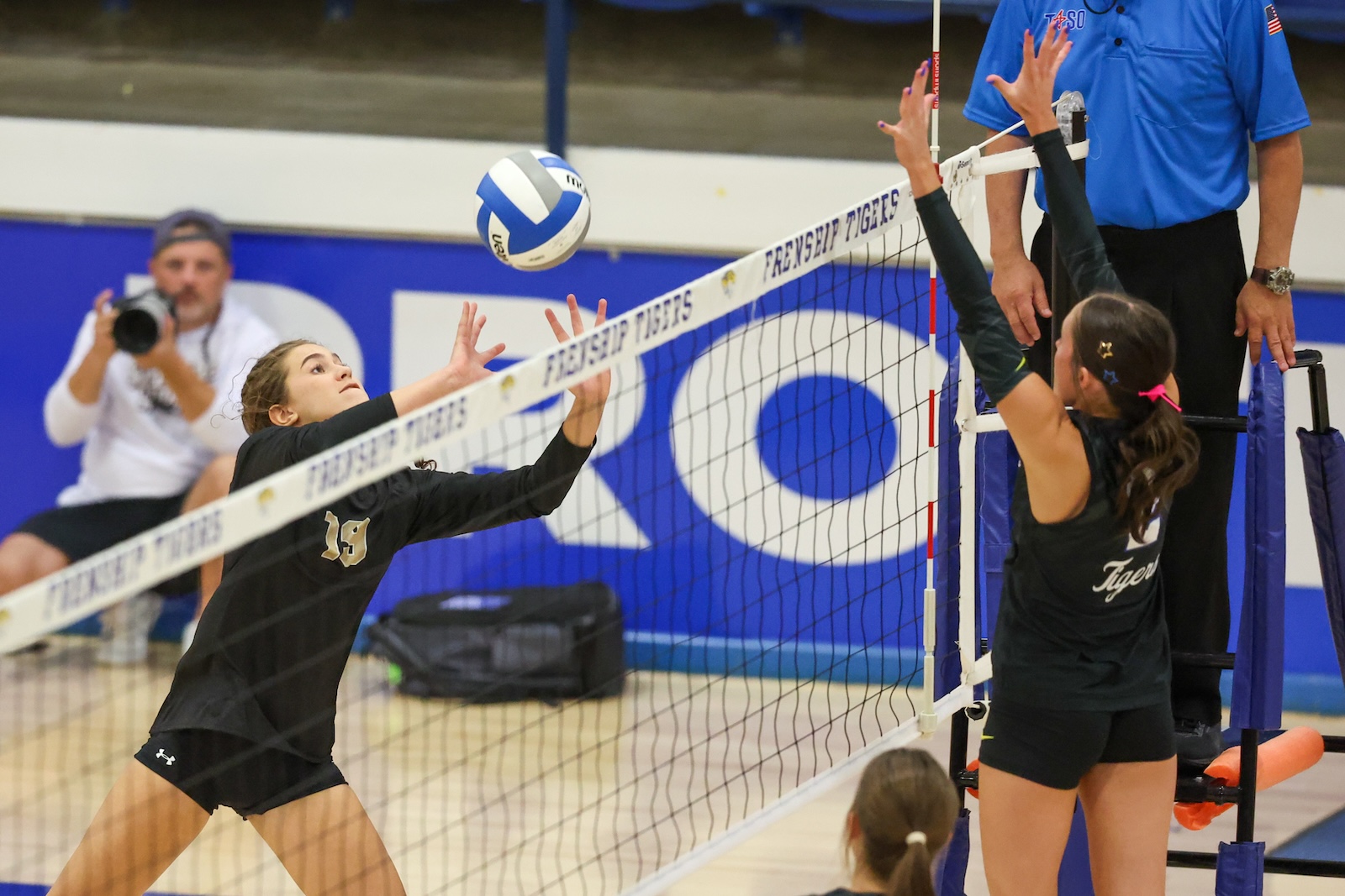 Seabolt Chase-13104 | Press Pass Sports A volleyball player in a black uniform dives to block the ball at the net as teammates watch, gym backdrop visible.
