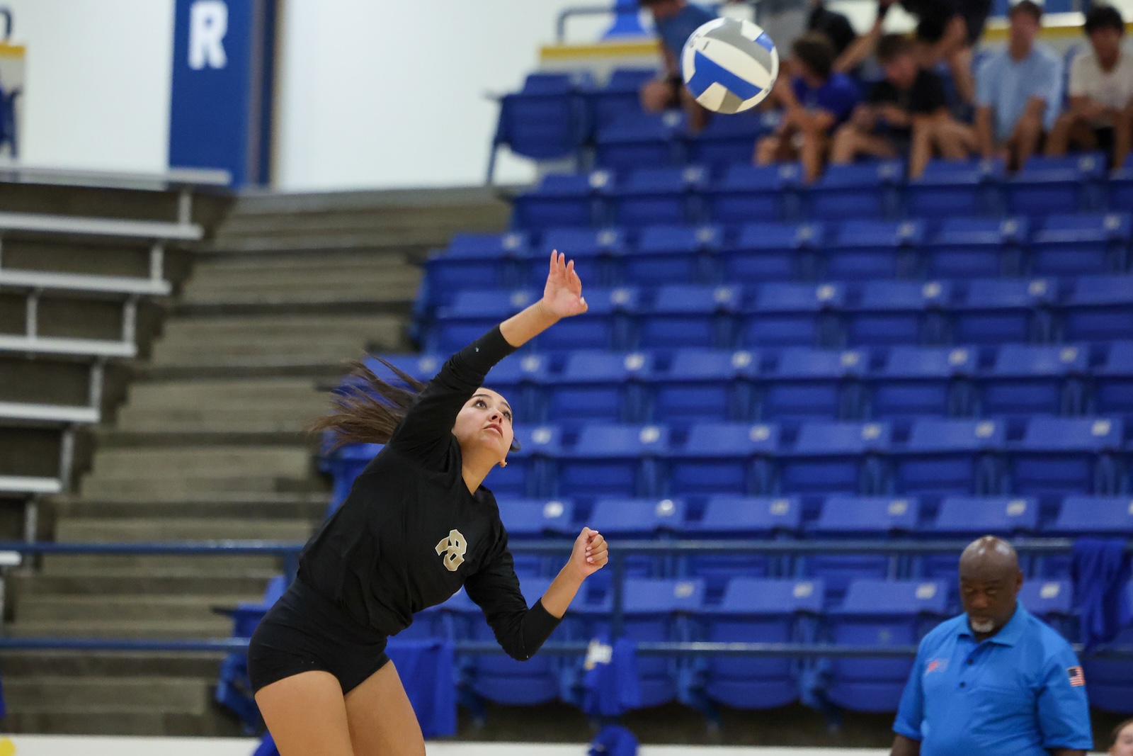Seabolt Chase-26052 | Press Pass Sports Female volleyball player in black reaching to strike a blue-and-white ball in an indoor gym; blue bleachers and spectators in the background
