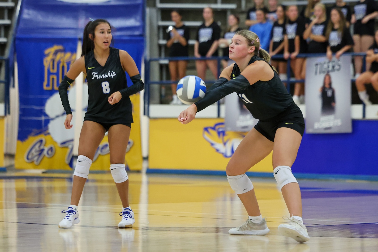 Seabolt Chase-26115 | Press Pass Sports Two volleyball players in black uniforms reach for a ball during a rally in a gym with spectators in the stands behind them, one wearing number 8 with white knee pads.