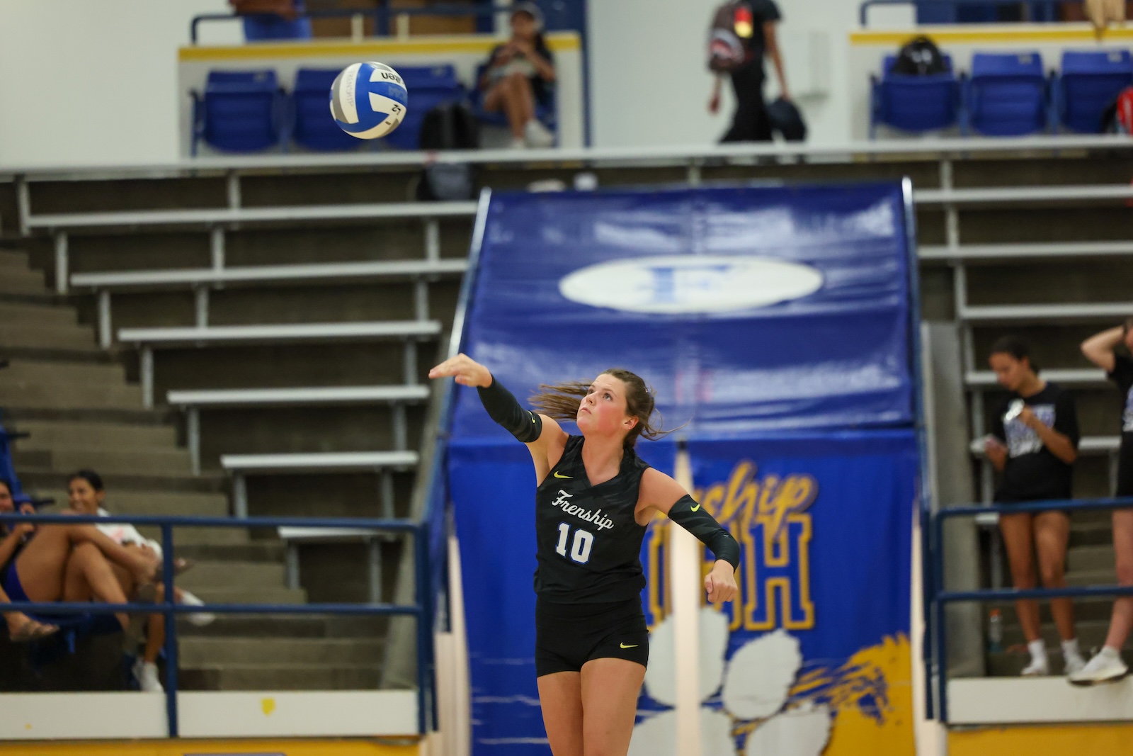 Seabolt Chase-26138 | Press Pass Sports Female volleyball player in a black jersey with number 10 serves the ball on an indoor court, blue bleachers in the background.