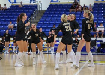 Seabolt Chase-26437 | Press Pass Sports Girls' volleyball team in black uniforms celebrate on a polished gym floor, blue bleachers in the background.