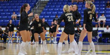 Girls' volleyball team in black uniforms celebrate on a polished gym floor, blue bleachers in the background.