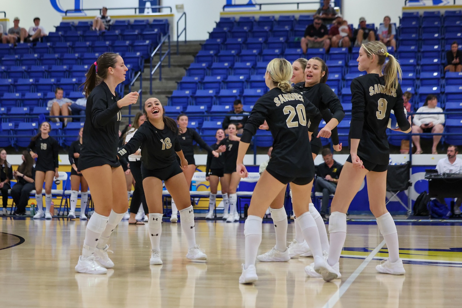 Seabolt Chase-26437 | Press Pass Sports Girls' volleyball team in black uniforms celebrate on a polished gym floor, blue bleachers in the background.