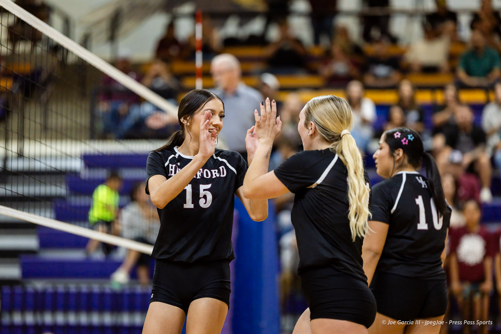 herford-vs-canyon-volleyball-jpegJoe-pps-1 | Press Pass Sports Two volleyball players in black uniforms high-five near the net during a match in a crowded gymnasium.