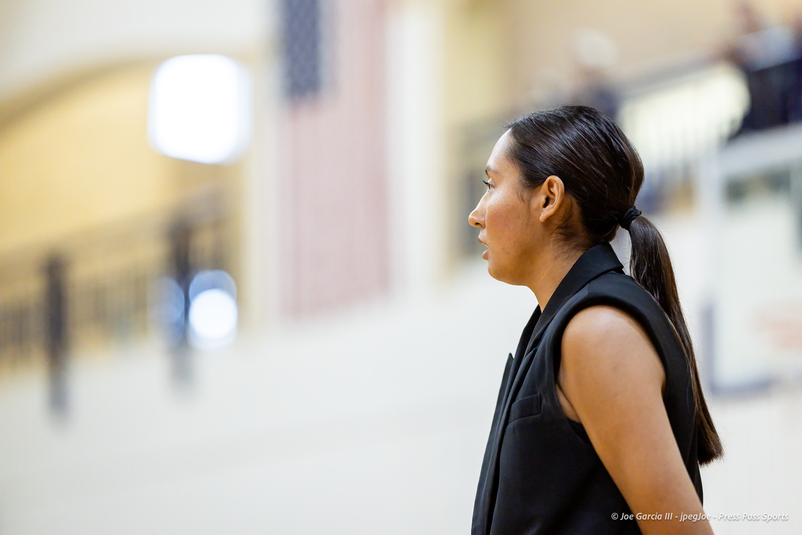 herford-vs-canyon-volleyball-jpegJoe-pps-4 | Press Pass Sports Side-profile of a woman with dark hair tied in a ponytail, wearing a black sleeveless jacket, standing in a bright indoor space with a blurred background