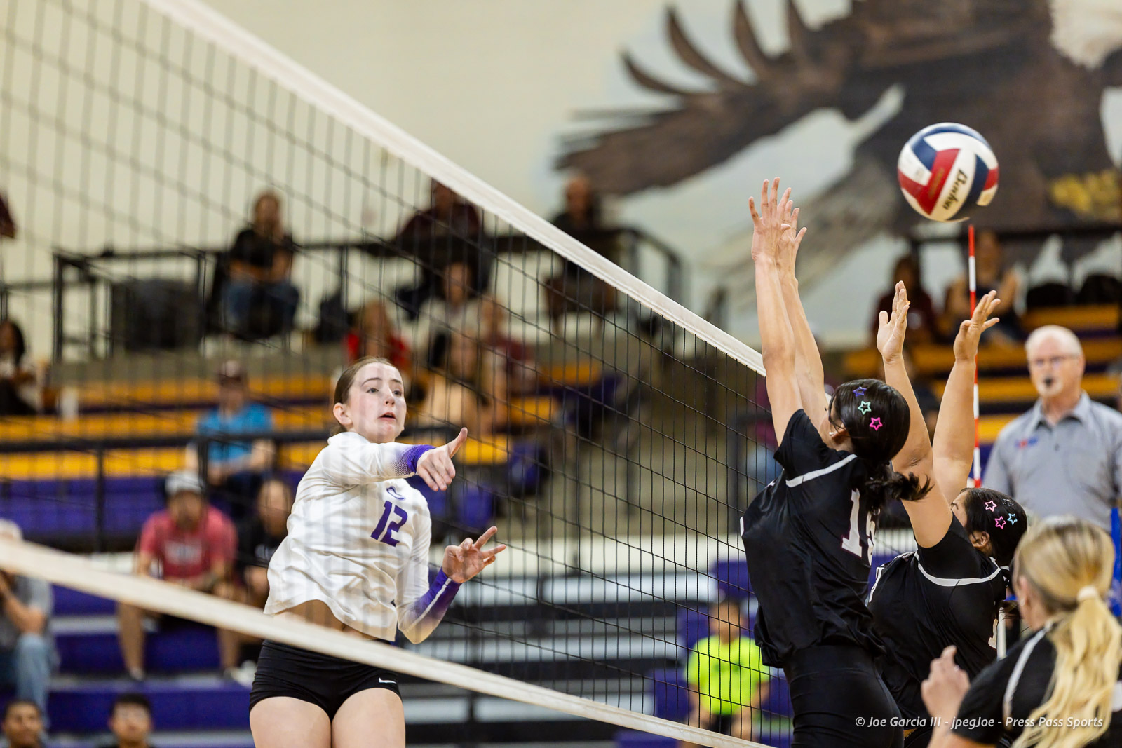 herford-vs-canyon-volleyball-jpegJoe-pps-5 | Press Pass Sports A volleyball player in a white jersey (#12) spikes the ball over the net while blockers in black jump to block it in a gym auditorium.