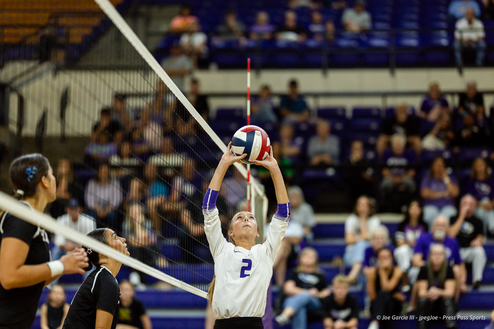 herford-vs-canyon-volleyball-jpegJoe-pps-12 | Press Pass Sports Female volleyball player wearing number 2 in a white jersey rises to set the ball over the net during a match, with teammates and a cheering crowd in the background.