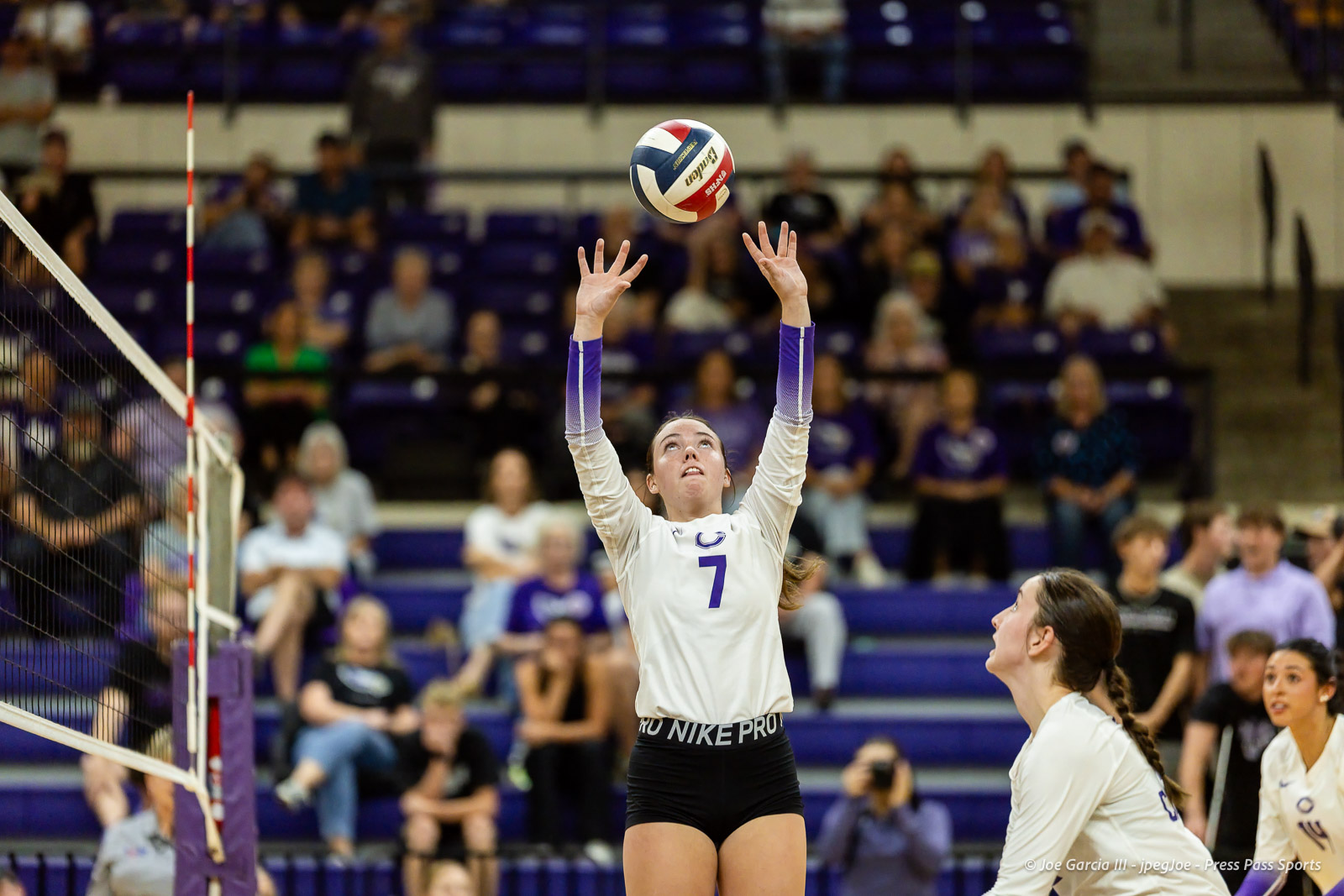 herford-vs-canyon-volleyball-jpegJoe-pps-13 | Press Pass Sports Female volleyball player in white jersey #7 jumps to set the ball over the net as spectators watch from the purple bleachers behind him/her.