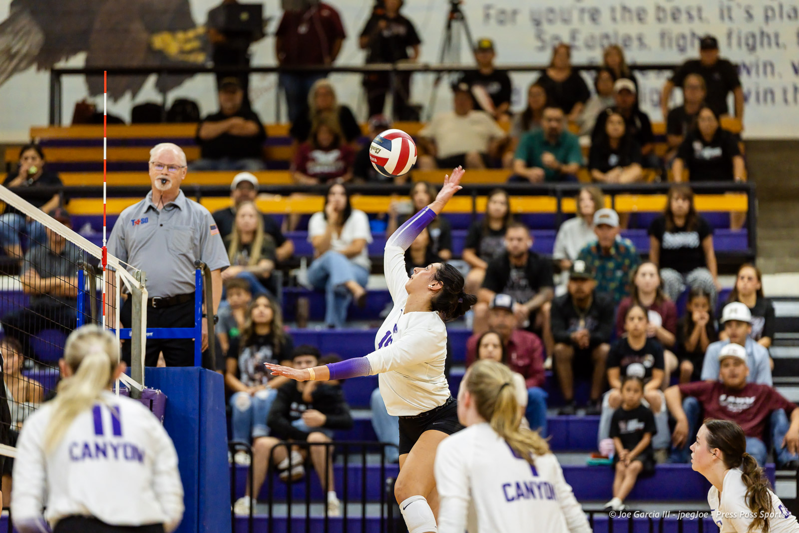 herford-vs-canyon-volleyball-jpegJoe-pps-20 | Press Pass Sports Female volleyball player in white reaching to hit the ball over the net during a match, with the referee and a crowded gym in the background.