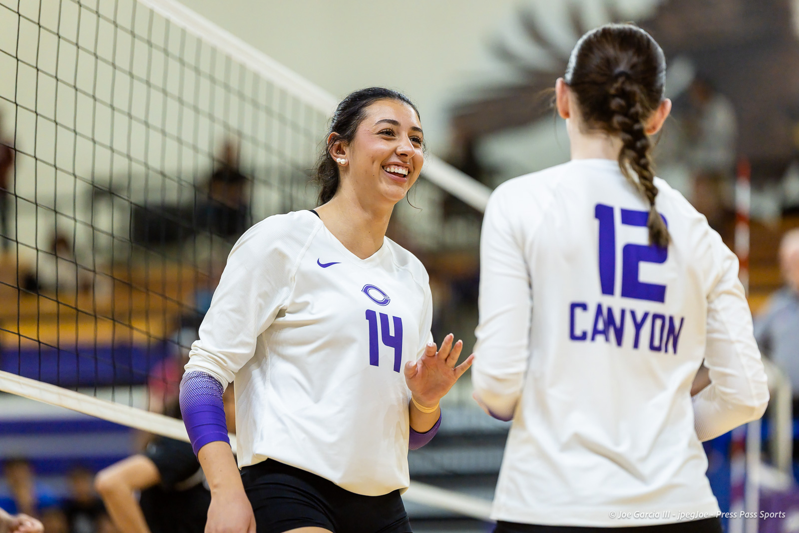 herford-vs-canyon-volleyball-jpegJoe-pps-23 | Press Pass Sports Two volleyball players in white uniforms share a smile at the net during a match, one facing the camera wearing #14 and a purple sleeve.
