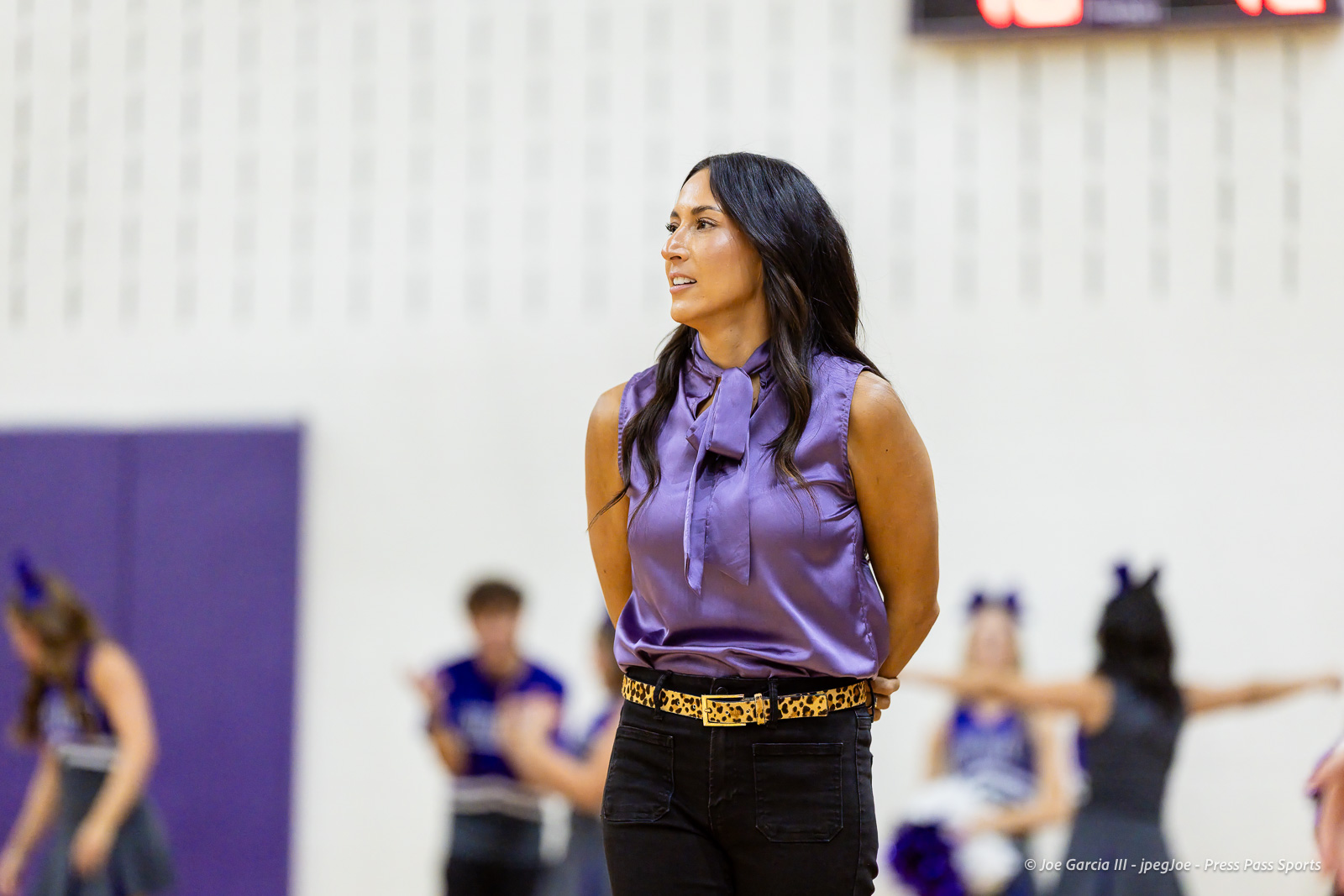 herford-vs-canyon-volleyball-jpegJoe-pps-24 | Press Pass Sports Dance instructor in a purple satin sleeveless blouse and leopard belt stands with hands behind back in a gym as dancers rehearse in the blurred background.