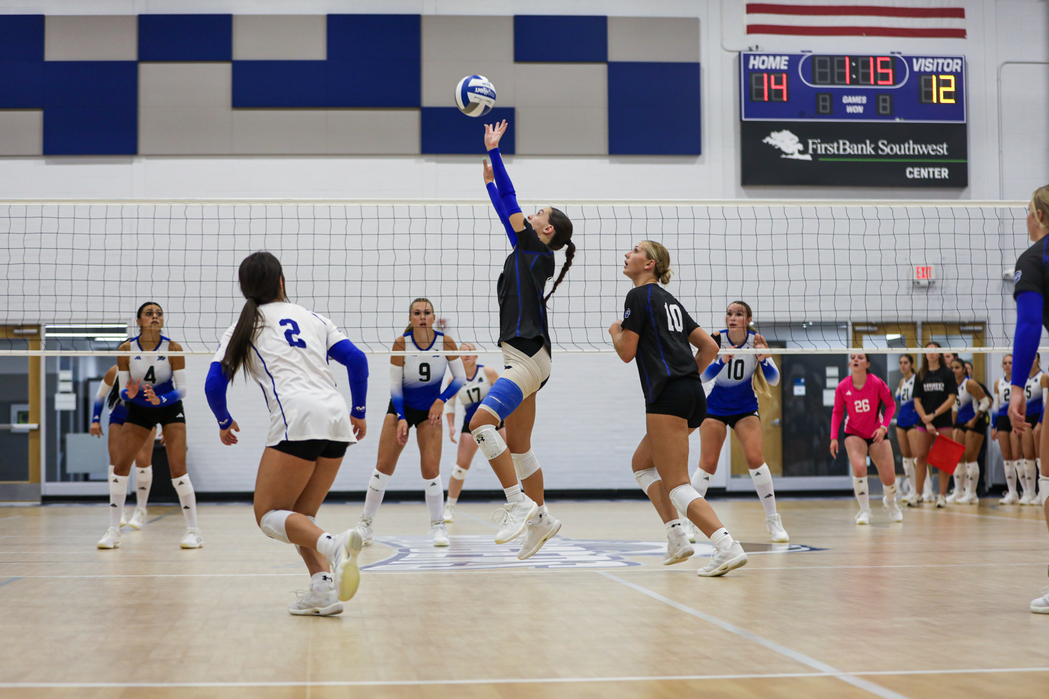 Edit 1 | Press Pass Sports Girls' varsity volleyball match: a player in blue sleeves jumps to hit the ball over the net while teammates watch from both sides in a gym.