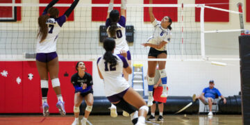 Hard spike at the net by a player in white jersey as opponents block at the net; teammates nearby on the court.