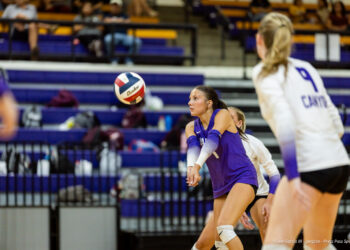 herford-vs-canyon-volleyball-jpegJoe-pps-19 | Press Pass Sports A female volleyball player in a purple uniform crouches with arms extended to pass a ball during a rally in an indoor gym, with teammates and spectators in the bleachers behind.