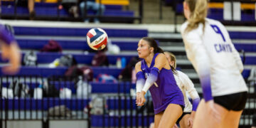 A female volleyball player in a purple uniform crouches with arms extended to pass a ball during a rally in an indoor gym, with teammates and spectators in the bleachers behind.