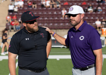 THS-CHS-FB-1 | Press Pass Sports Two coaches on a football field converse, one in black with a Texas Tech cap and sunglasses, the other in purple with a white Cincinnati cap.