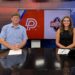 IMG_7328 | Press Pass Sports Two news anchors sit at a desk in a television studio, smiling, with a large screen displaying red and logo panels behind them to the left and right.