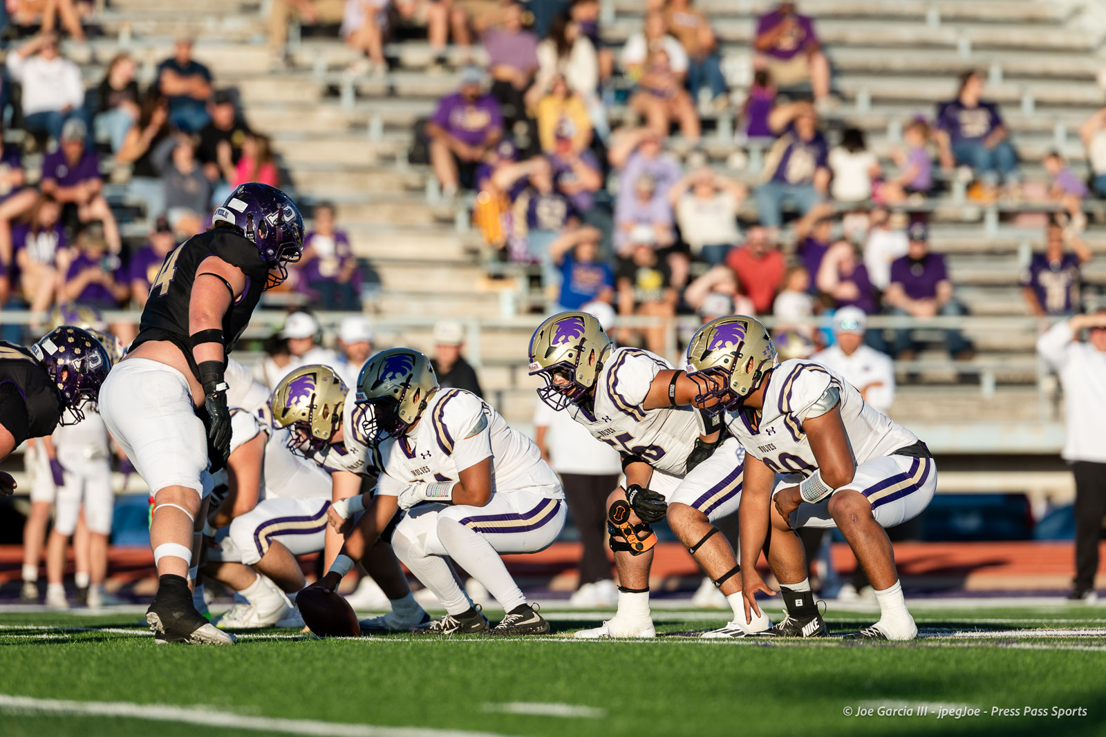 dalhart-vs-panhandle-football-jpegJoe-pps-6 | Press Pass Sports Football players line up at the line of scrimmage during a game; white and purple uniforms, crowd in bleachers behind.