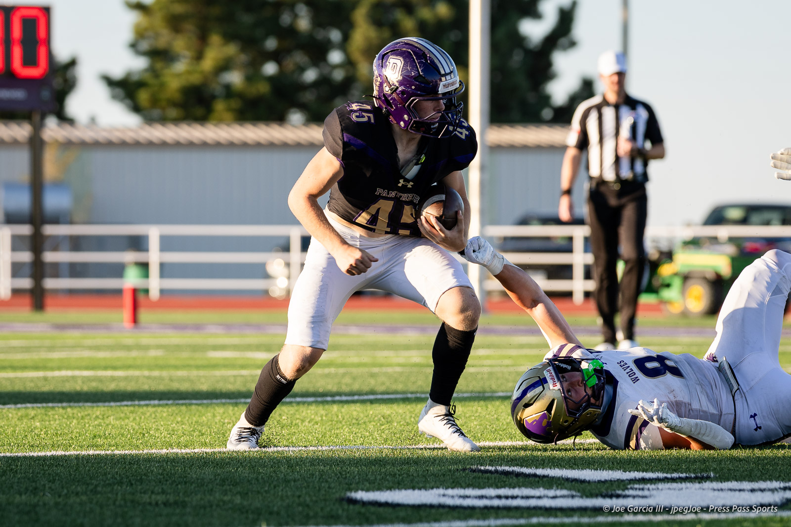 dalhart-vs-panhandle-football-jpegJoe-pps-8 | Press Pass Sports Football player in purple and black jersey runs with the ball as a defender on the ground reaches for him; referee visible in the background on a green field.