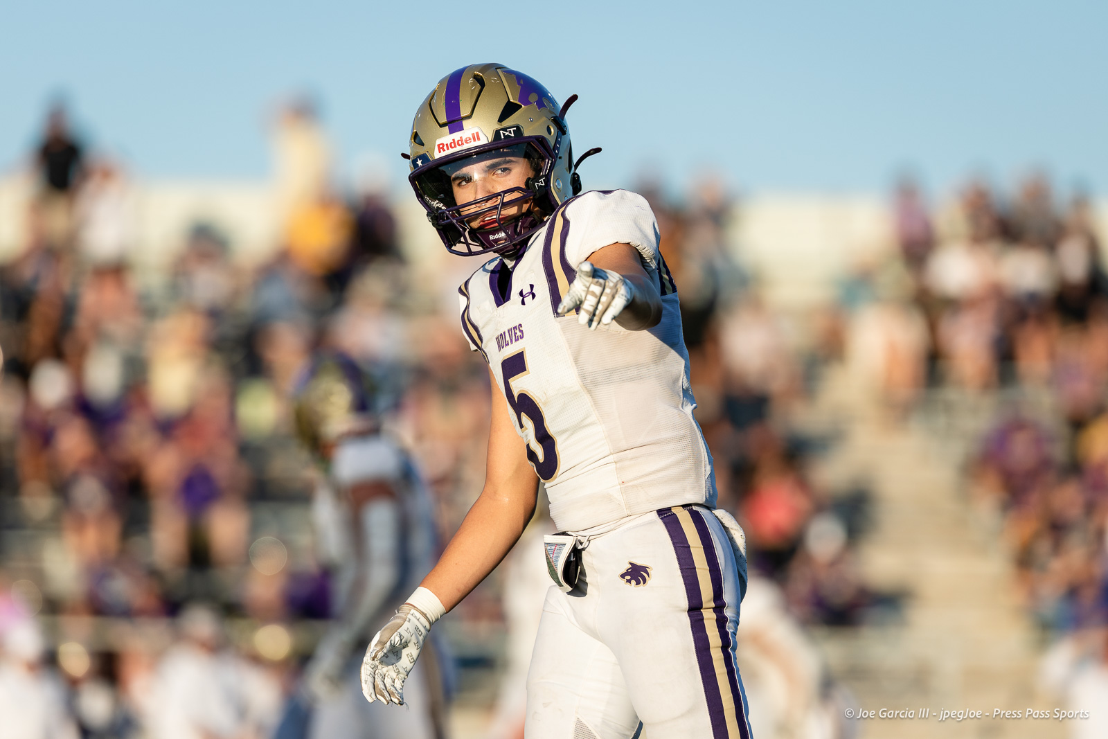dalhart-vs-panhandle-football-jpegJoe-pps-10 | Press Pass Sports Football player in white and purple uniform pointing toward the camera on a sunny field with a blurred crowd in the stands behind him.