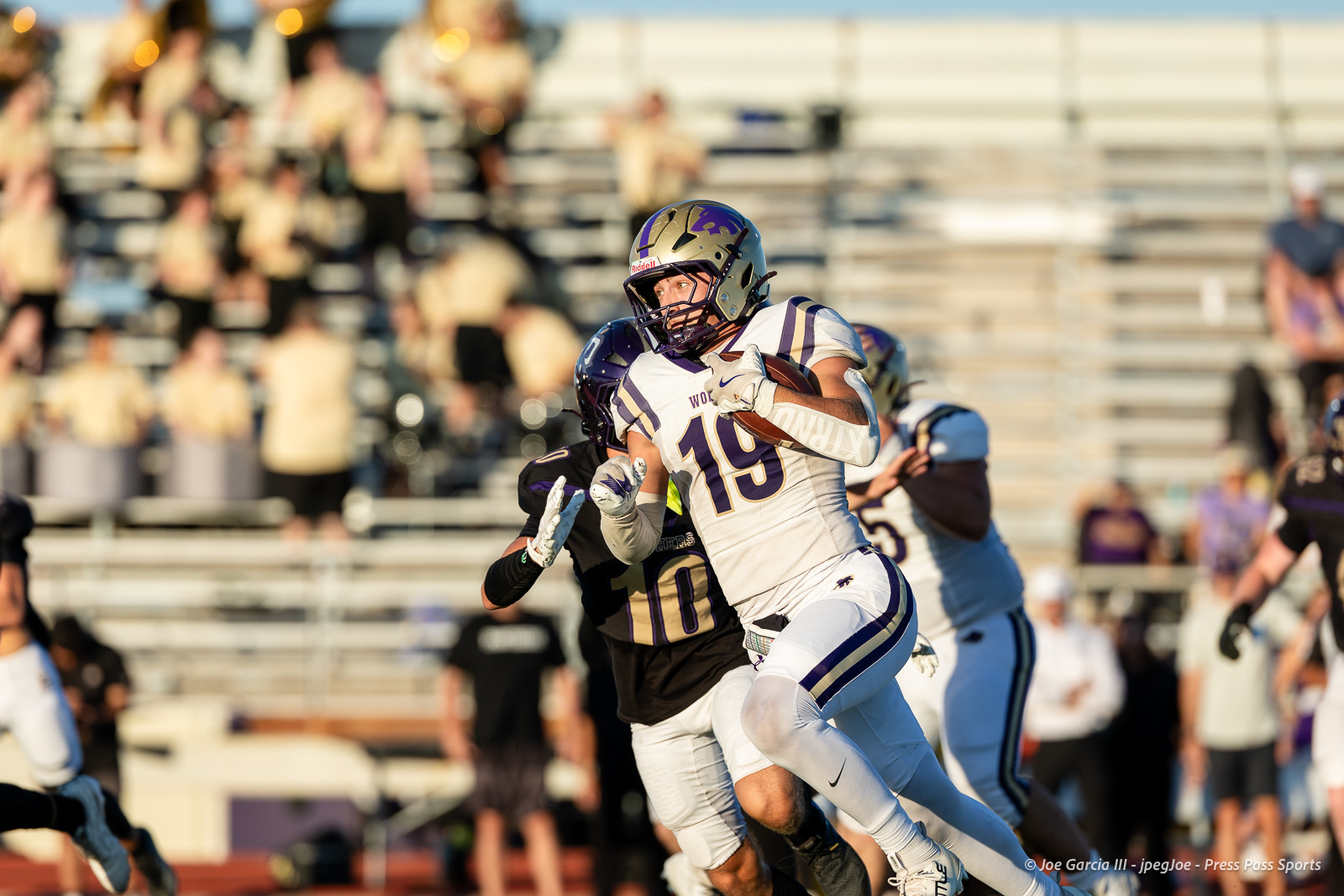 dalhart-vs-panhandle-football-jpegJoe-pps-12 | Press Pass Sports Football player in white and purple jersey #19 carries the ball and runs downfield during a game, with a defender close behind in a stadium crowd backdrop.