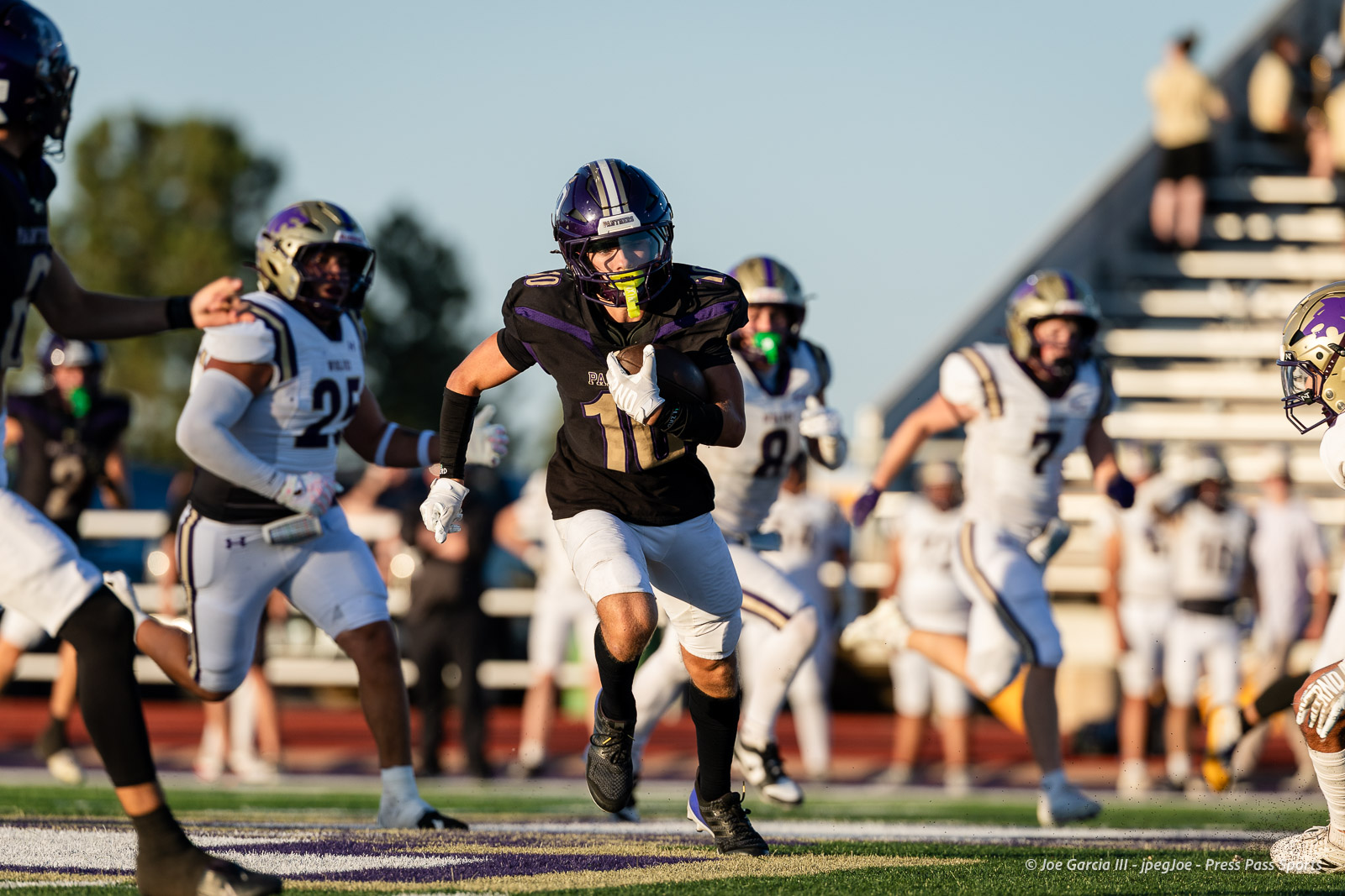dalhart-vs-panhandle-football-jpegJoe-pps-13 | Press Pass Sports Football player in black and purple uniform runs with the ball, pursued by white-uniformed defenders on a sunny field.