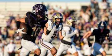 Football player wearing black and purple Panthers uniform with the ball racing down the field as defenders pursue, stands full of spectators in the background.