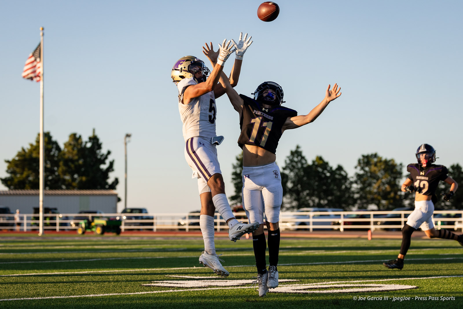 dalhart-vs-panhandle-football-jpegJoe-pps-15 | Press Pass Sports Two football players leap for a pass on a sunny field, with an American flag and bleachers in the background.