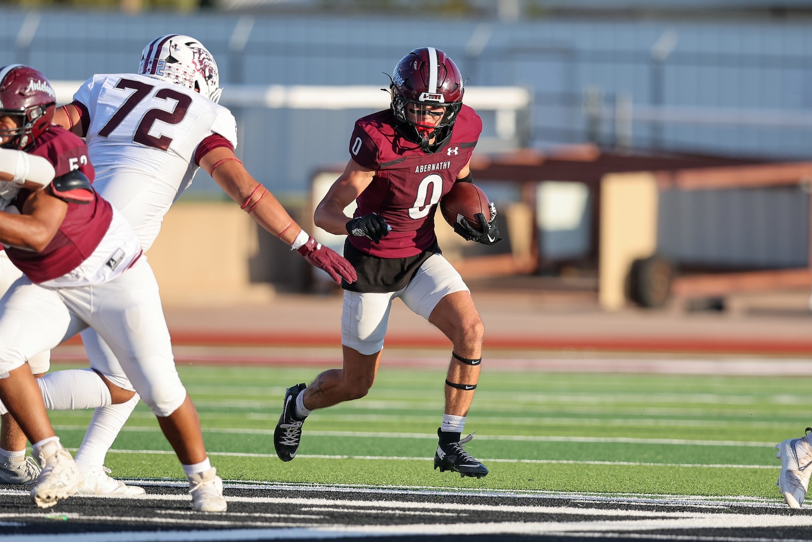 Football player in maroon Abernathy jersey with the ball runs forward as white-clad defenders pursue.