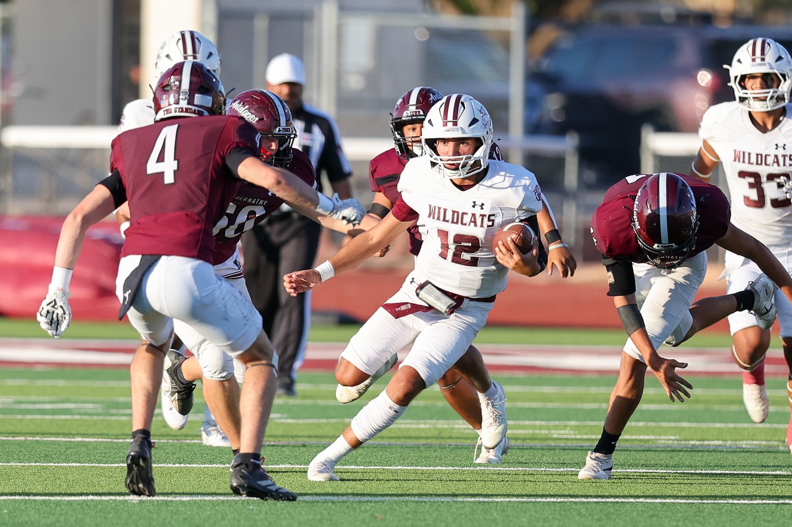 Football action: a Wildcats player in white #12 carries the ball while maroon-clad players close in on a green turf field.