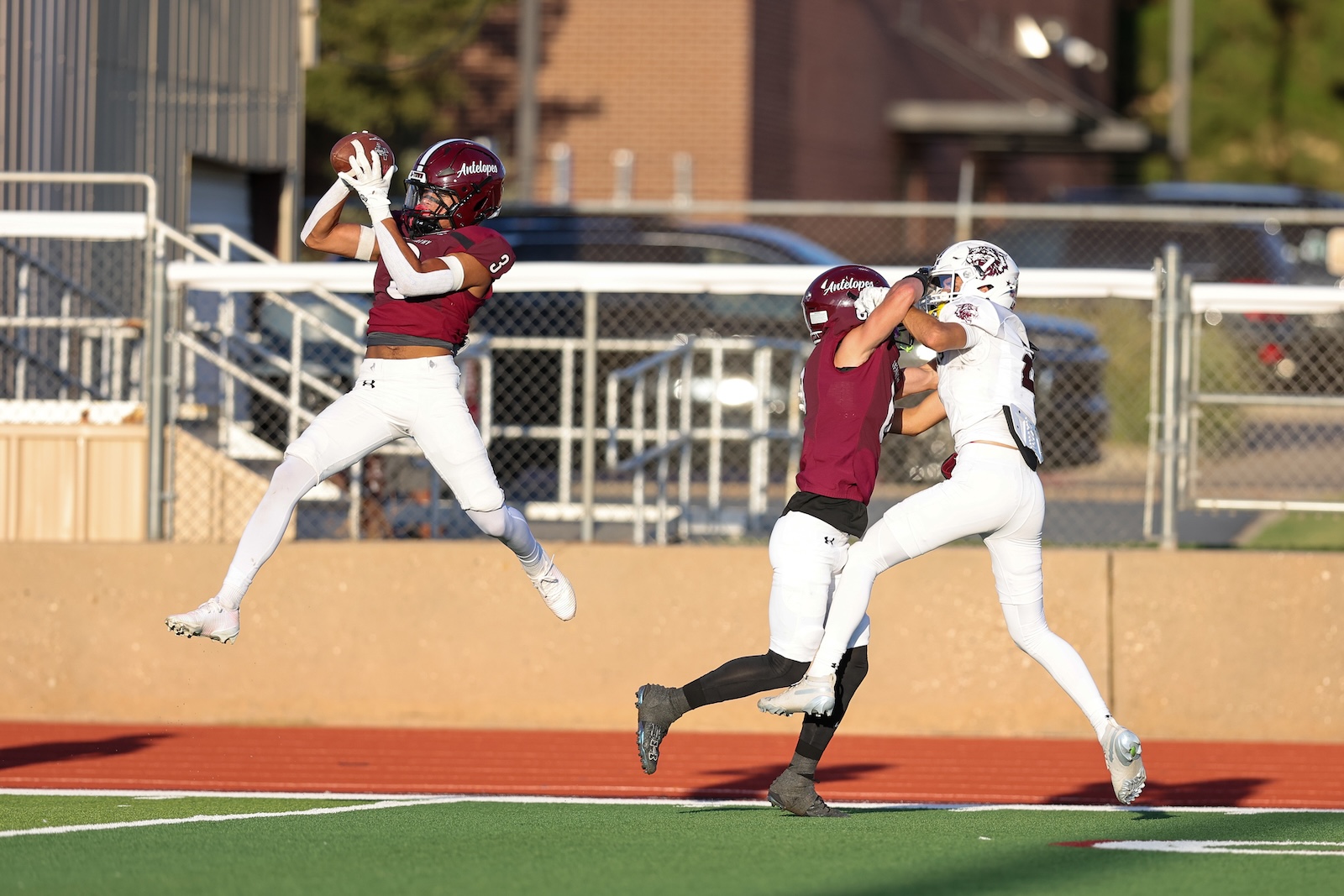 A maroon-jersey receiver leaps to catch a football while a defender in white blocks nearby on a football field under a clear sky.