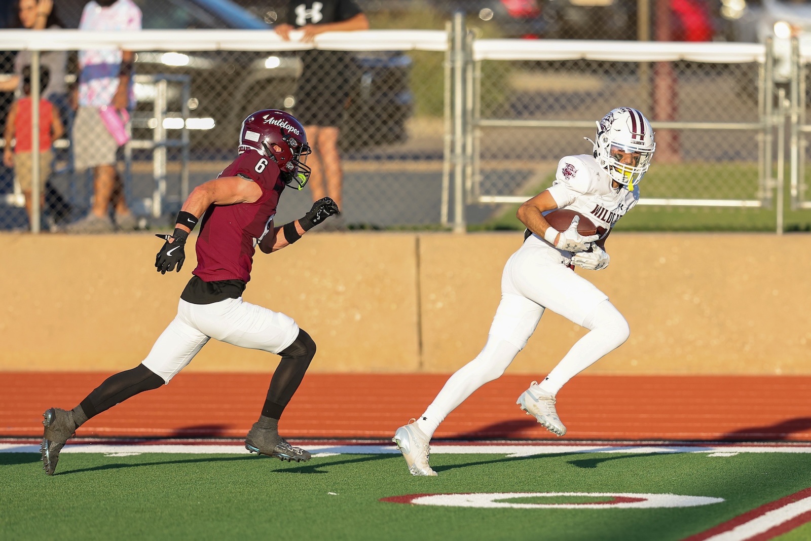 A football player in maroon jersey chases a ball carrier in white on a turf field during a game with spectators in the background.