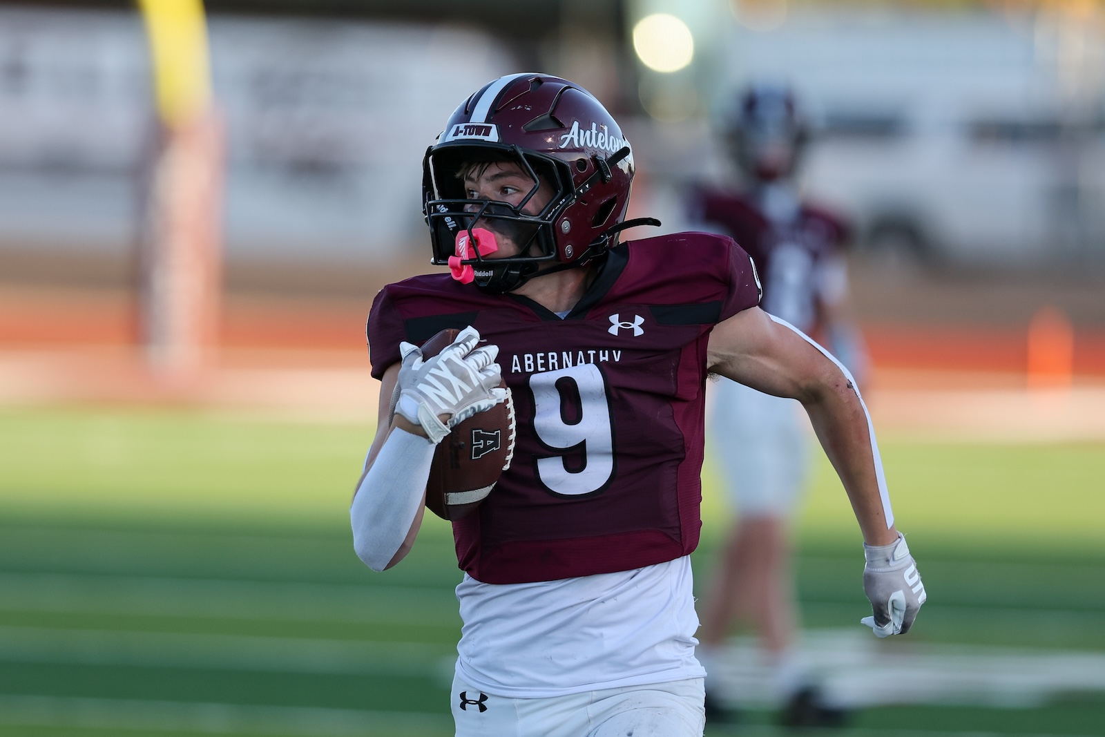 Football player in maroon jersey #9 runs with the ball on a green field, Abernathy team.