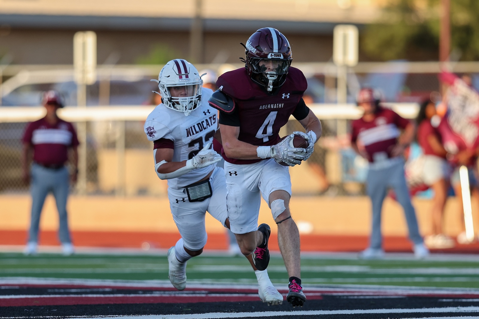 Football players in a maroon Abernathy uniform and a white Wildcats uniform sprint on a turf field, pursuing the ball carrier in a daytime game.