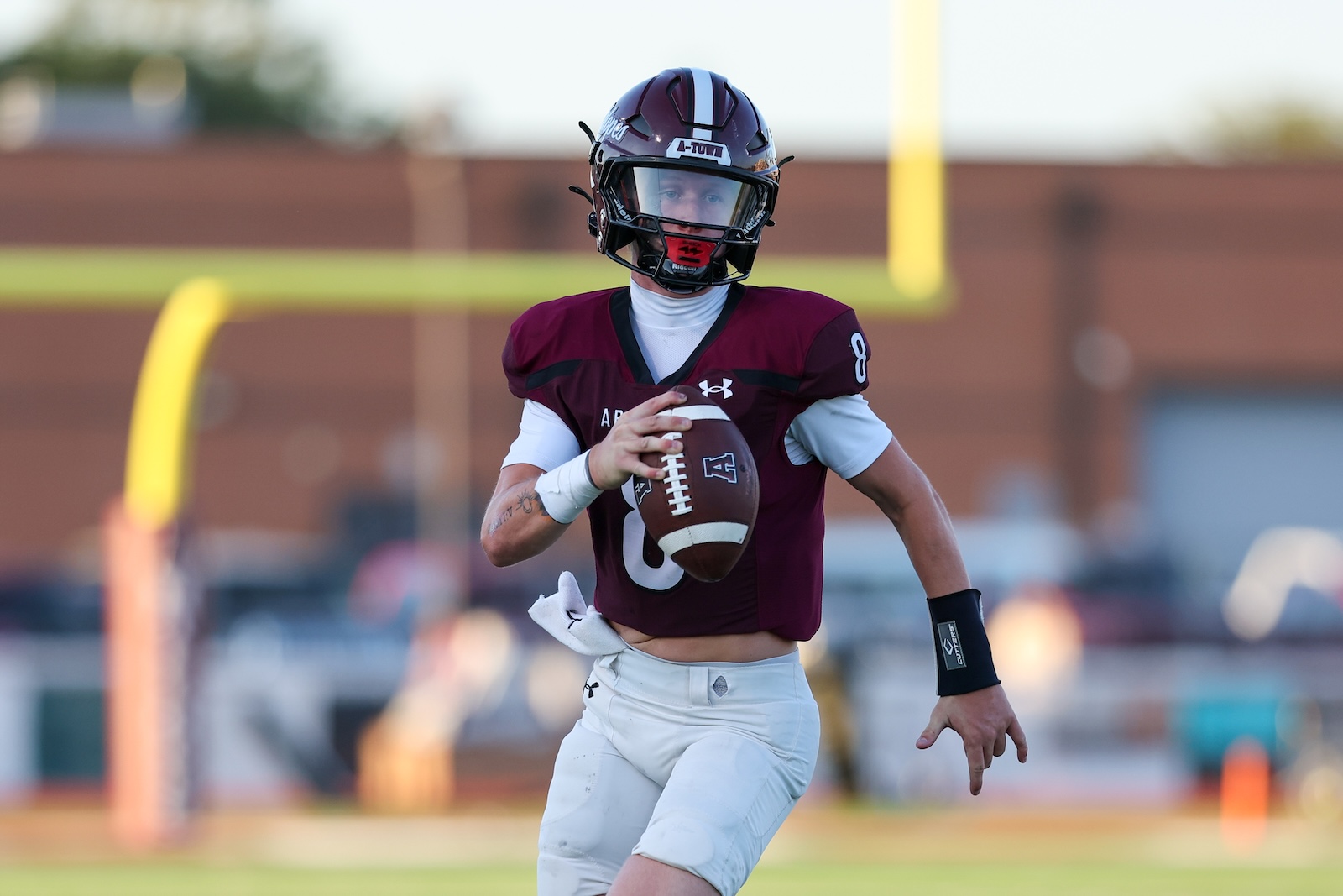 Football player in maroon jersey runs with the ball on a field, helmet on, during a game