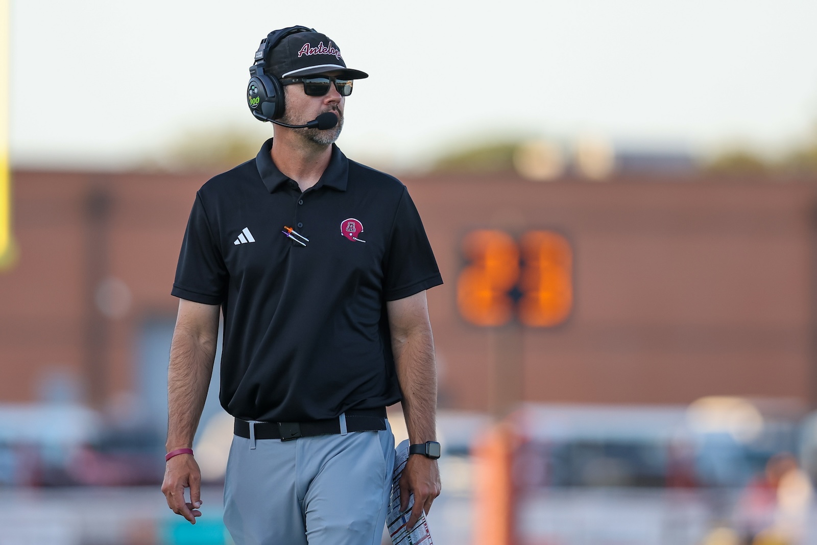 Coach wearing a headset and sunglasses on the football sideline, holding a towel or clipboard.