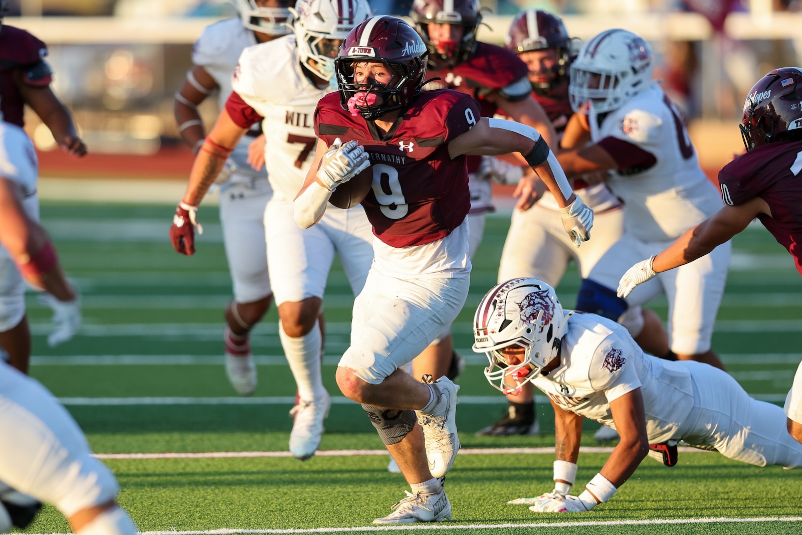 Football player in maroon #9 carries the ball as defenders chase on a sunny field.