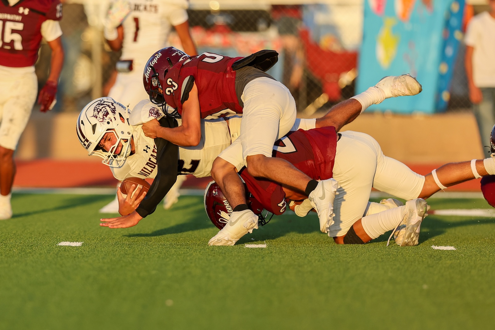 A football player in white with a Wolves logo is tackled by a tackler in maroon on a green turf field during a game in progress.
