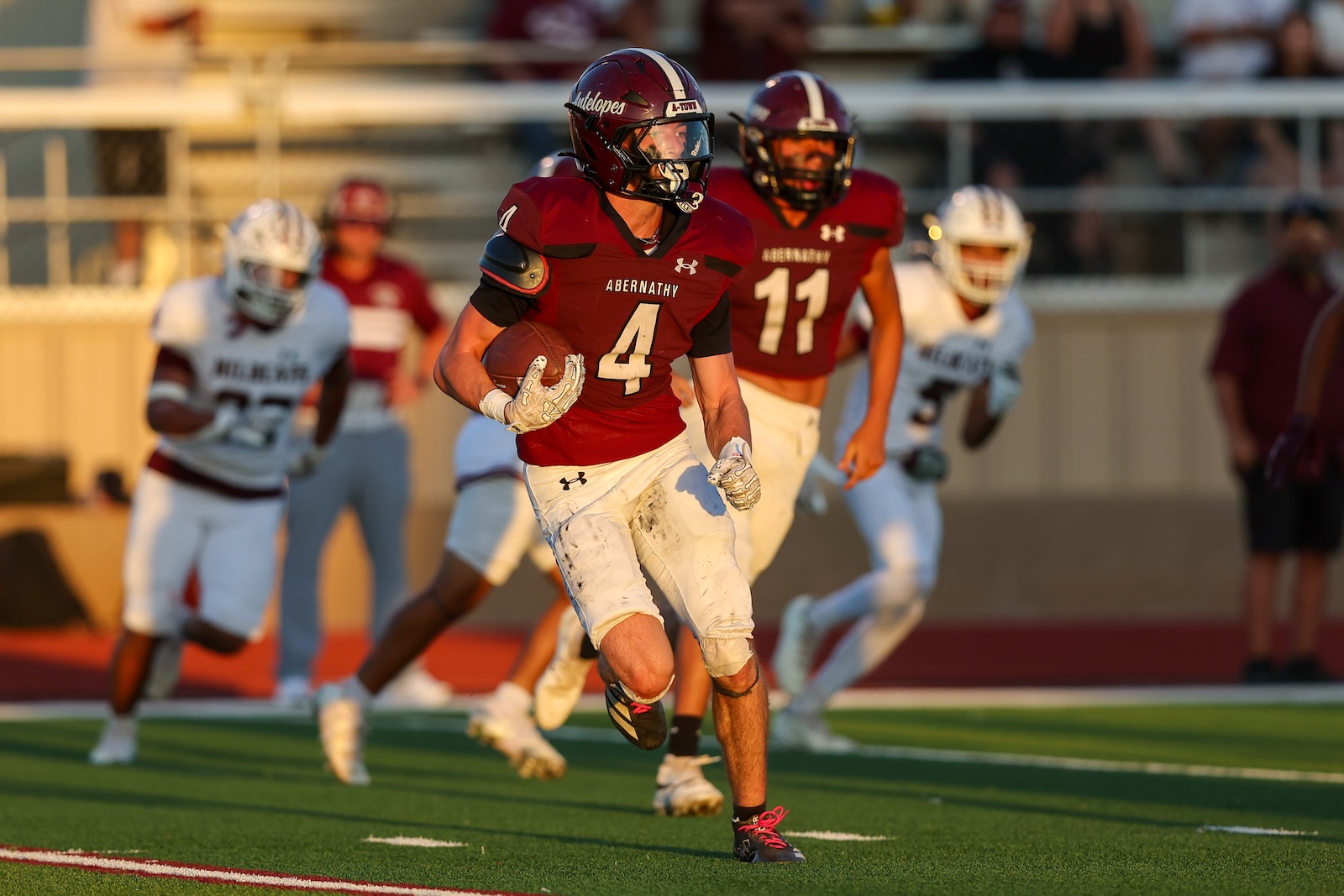 Abernathy #4 football player in maroon jersey runs with the ball on a green field, pursued by teammates in the background.