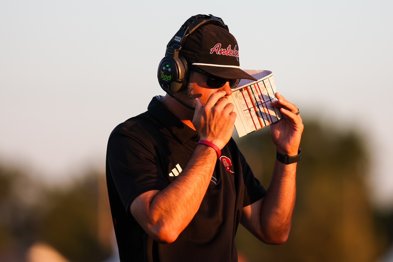 Man wearing a headset and sunglasses reads a playbook up to his mouth on a sunny outdoor field, wearing a black polo and a cap with red lettering.
