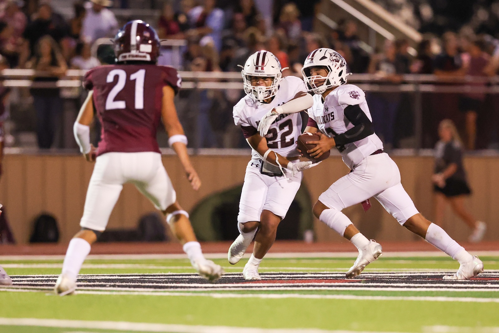 Two football players in white jerseys tackle a ball carrier while a maroon-clad defender (#21) pursues in a stadium.